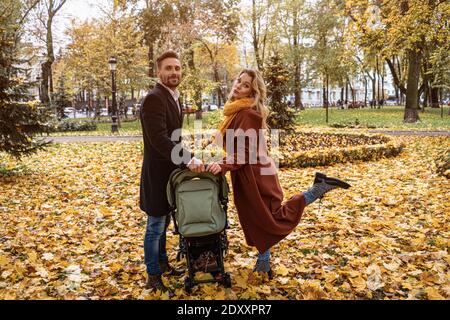 Glückliche Familie mit einem neugeborenen Baby im Kinderwagen zu Fuß in einem Herbstpark. Glückliche Mutter posiert einen Kinderwagen mit einem Bein im Freien in einem goldenen halten Stockfoto