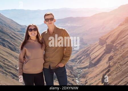 Aufgeregt Paar in der Liebe Umarmungen und lächelt gegen große stehen Felsige braune Berge mit Schlucht und kleinen Fluss unter hell Herbstsonne Stockfoto