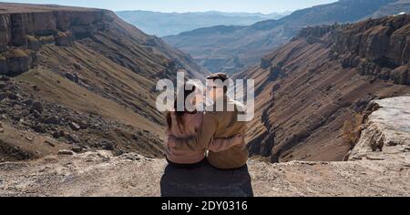 Panorama von romantischen Paar in der Liebe Umarmungen und Blicke in Augen tragen eine Sonnenbrille und sitzen auf einer steilen Klippe gegen Berglandschaft Stockfoto