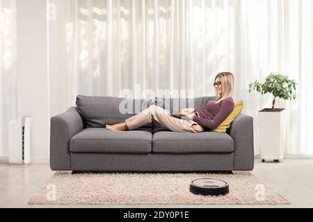 Junge Frau, die auf einem Laptop arbeitet und auf einem liegt Sofa zu Hause mit einem Roboter-Staubsauger auf dem Teppich Stockfoto