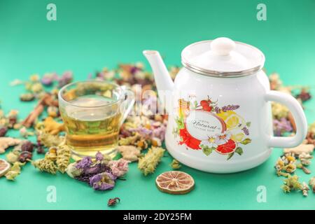 Kräutertee in Teekanne und Glas Tasse auf grünem Hintergrund unter trockenen Kräutern Salbei Hagebutte Kurkuma Kamille, selektiver Fokus. Stockfoto