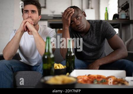 Unzufrieden multikulturelle Männer in Casual Outfit klammend Hände und berühren Stirn beim Sitzen auf Sofa neben Tisch mit Snacks und Bier und beobachten Stockfoto