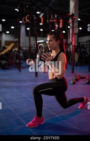 Seitenansicht einer starken Sportlerin, die atmet und Ausfallschritte macht Mit schwerer Kettlebell beim Funktionstraining im modernen Fitnessstudio Stockfoto