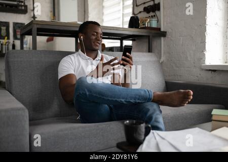 Positiver ethnischer Kerl in legerer Kleidung, der auf dem Sofa sitzt und Videoanruf mit Kommilitonen während des Studiums zu Hause Stockfoto