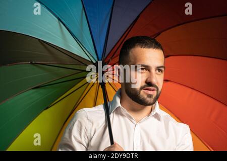 Bärtiger Kerl halten bunten Regenschirm. Ein netter Kerl mit einem stilvollen Bart und weißem Hemd steht mit einem Regenbogenschirm hinter seinem Rücken. Gut aussehender Kerl Stockfoto