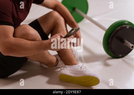 Body Nahaufnahme eines jungen Mannes mit Smartphone in der Hand Surfen im Internet oder SMS auf dem Boden sitzen, schwarz und grün Ton Fitness Langhantel, Geräte Stockfoto