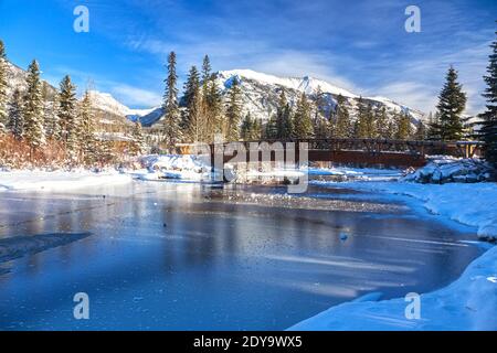 Fußgängerbrücke aus Holz über dem Frozen River Ice. Die schneebedeckte Rocky Mountain Peaks Blue Skyline. Kanadische Rockies – Winterlandschaft Banff National Park, Alberta Stockfoto