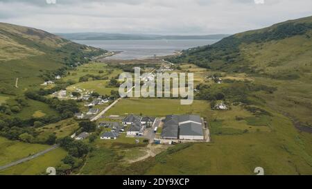 Schottische Whiskybrennerei in Village Aerial. Straße, Ferienhäuser, Häuser im grünen Bergtal. Sommer bewölkt Tag. Niemand Natur Landschaft von Arran Island, Schottland. Drohnenaufnahme im Kino Stockfoto