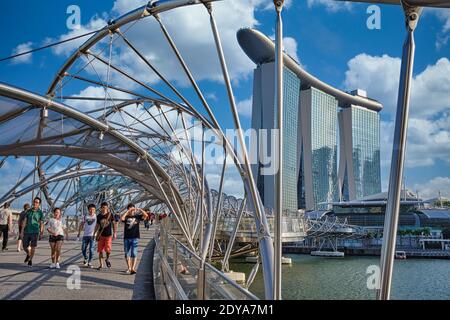 Touristen, die über die futuristisch anmutende Helix Bridge mit dem ikonischen Marina Bay Sands Hotel im Hintergrund fahren; Marina Bay, Singapur Stockfoto