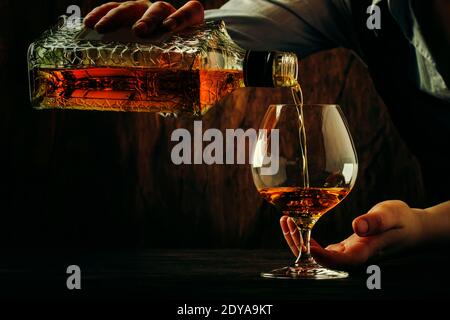 Der Barkeeper gießt den Cognac oder Brandy in ein großes Weinglas an der alten Theke. Vintage Holzhintergrund in Pub oder Bar, Nachtstimmung. Für Text platzieren Stockfoto