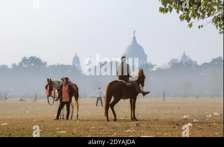 Einheimische Reiter mit ihren Pferden warten auf Touristen in Kalkutta Maidan Bereich mit Blick auf Victoria Memorial Denkmal auf einem Nebliger Wintermorgen Stockfoto