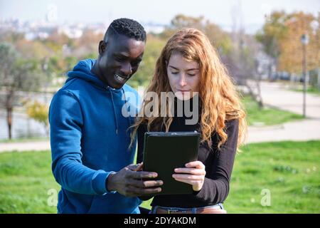 Afrikanischer schwarzer Mann und Rotschopf kaukasische Frau, die ihre Tablette in einem Park betrachten. Junges multirassisches Paar. Stockfoto
