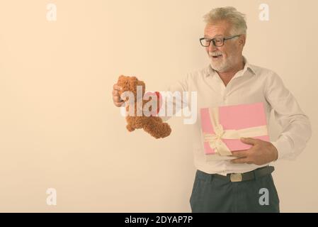 Studioaufnahme eines glücklichen älteren bärtigen Mannes, der lächelt, während er hält Geschenkbox und Blick auf Teddybär mit Herz und Liebeszeichen beim Tragen von Brillen Stockfoto