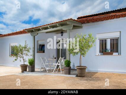 Moderne Fassade des weißen Wohnhauses mit rotem Fliesendach, Topfpflanzen, Essmöbel im Hinterhof in der Nähe von Glastür Gebäudeeingang Stockfoto
