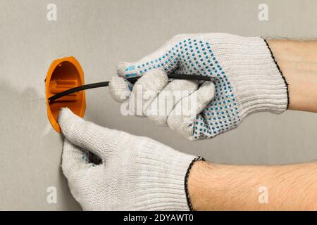 Die Hand eines Elektrikers in einem Handschuh leitet das Kabel in die Kastenbuchsen an der Trockenwand, in der Nähe Stockfoto