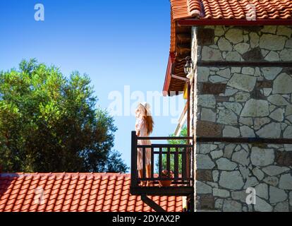 Seitenansicht der jungen Frau, die auf dem Holzbalkon steht Altes Kopfsteinpflasterhaus mit rotem Ziegeldach im Sommer Landschaft auf dem Land Stockfoto