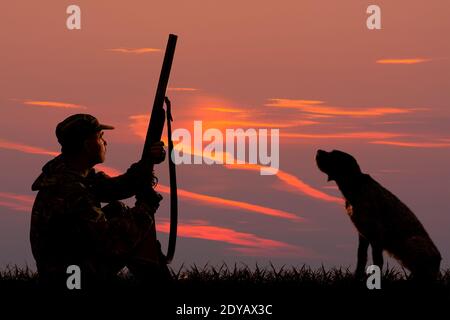 Silhouetten eines Jägers und eines Hundes, der sitzt Bei Sonnenuntergang Hintergrund auf der Jagd Stockfoto