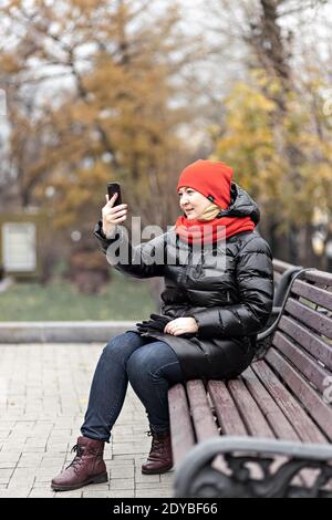 Eine junge Frau kommuniziert im Online-Video-Chat auf einem Handy, sitzt auf einer Bank in einem Herbstpark. Soziale Distanzierung und Isolation während des CO Stockfoto