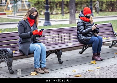 Zwei junge Frauen mit medizinischen Masken sprechen auf einem Handy in sicherer sozialer Distanz, während sie auf einer Bank in einem Herbstpark sitzen. Meeting und chatti Stockfoto