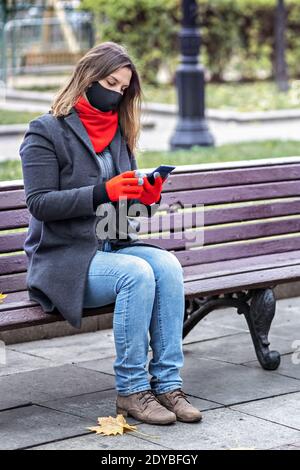 Eine junge Frau in einer medizinischen Maske kommuniziert online chattet auf einem Handy, sitzt auf einer Bank in einem Herbstpark. Soziale Distanzierung und Isolation Stockfoto