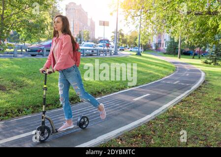 Mädchen im Sommer in der Stadt reitet einen Roller, in lässiger Kleidung Jeans und eine rosa Jacke, freien Platz für eine Kopie des Textes. Hintergrund Straße grünes Gras und Stockfoto