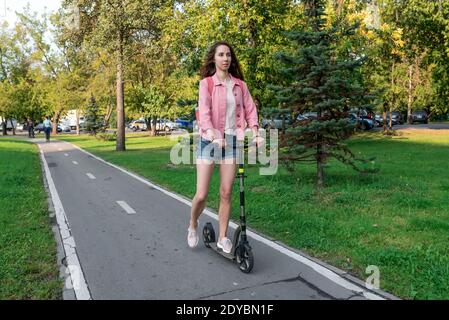 Frau im Sommer in der Stadt fährt Roller auf einer Fahrradstrecke, in legerer Kleidung in Jeans-Shorts und einer rosa Jacke, freier Platz für Kopie des Textes. Hintergrund Stockfoto