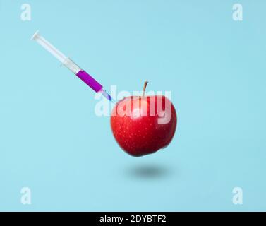 Genetisch modifizierter roter Apfel mit Spritzen auf blauem Hintergrund. Medizinische Spritze, die GVO in Apfel injiziert, Fruchtkonzept minimal Stockfoto