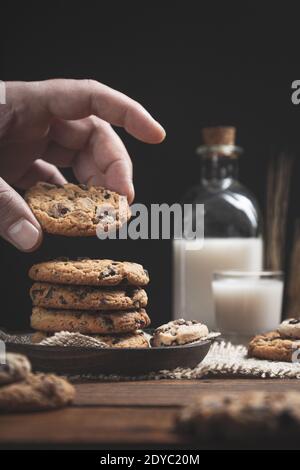 Hand hält Schokoladenkekse mit einem Glas und einer Flasche Milch auf einem Holzsockel, dunkler Hintergrund. Konzept für süße Speisen. Stockfoto