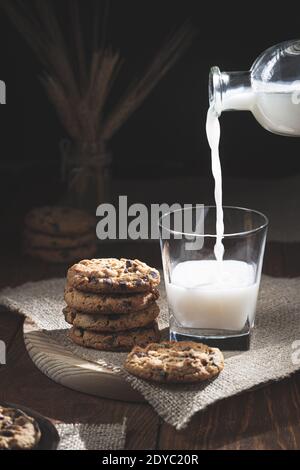 Chocolate Chip Cookies und Milchflasche verschüttet Milch in einem Glas auf einem Holzsockel, dunkler Hintergrund. Konzept für süße Speisen. Stockfoto