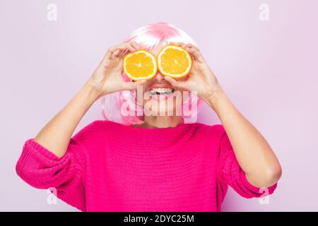 Kaukasische Frau mit rosa Perücke und Pullover, mit zwei orangefarbenen Scheiben in den Augen posiert lustig im Fotostudio. Lustige ausdrucksstarke Frauen und bunte po Stockfoto