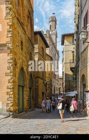 Blick auf die Altstadt von Volterra mit Touristen zu Fuß in einer engen Straße mit dem mittelalterlichen Palazzo dei Priori im Hintergrund, Toskana, Italien Stockfoto