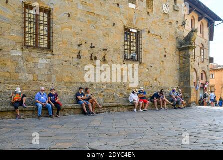 Touristen mit Covid-19 Masken sitzen auf einer langen Steinbank vor dem Palazzo dei Priori in der Altstadt von Volterra, Pisa, Toskana, Italien Stockfoto