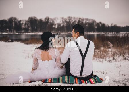 Mexikanische Braut und Bräutigam in Hochzeitskleidung und Hosenträger sitzen auf dem Schnee im Winter. Frau in Kleid und schwarzen Hut Stockfoto
