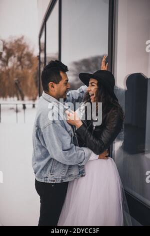 Mexikanische hispanischen Mann und Frau im Hut küssen Umarmung lachend zu Fuß. Bräutigam Braut Hochzeit Liebe Paar im Winter im Schnee auf dem Hintergrund von Shop wi Stockfoto