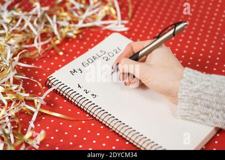 Weibliche Hände schreiben meine Ziele 2021 in einem Notizbuch. Roter Hintergrund und Lametta. Konzept für das neue Jahr. Stockfoto