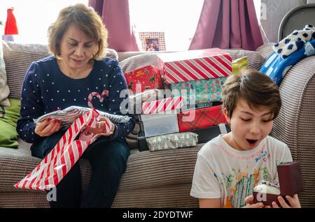 Ein Junge ist aufgeregt und überrascht, dass er am Weihnachtstag ein Weihnachtsgeschenk geöffnet hat, während seine Großmutter neben ihm Geschenke öffnet. Stockfoto