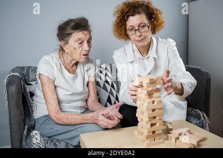 Jenga-Spiel. Thema ist Demenz, Altern und Spiele für alte Menschen. Kaukasische ältere Frau baut Turm aus Holzblöcken mit Hilfe eines Arztes als Teil Stockfoto