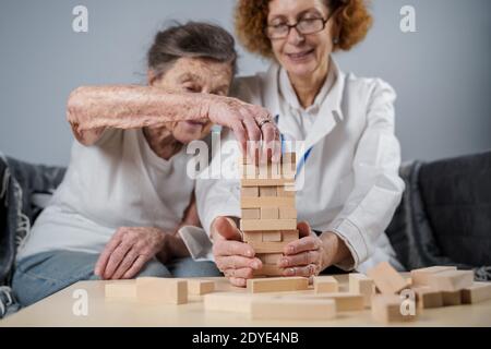 Jenga-Spiel. Thema ist Demenz, Altern und Spiele für alte Menschen. Kaukasische ältere Frau baut Turm aus Holzblöcken mit Hilfe eines Arztes als Teil Stockfoto