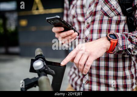 Männliche Hände modernen Elektroroller schaltet sich ein und Melodien. Kaukasischen Mann gekleidet stilvoll lässig verwendet Handy, um Kick Scooter zu steuern, close-up nein Stockfoto