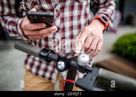 Männliche Hände modernen Elektroroller schaltet sich ein und Melodien. Kaukasischen Mann gekleidet stilvoll lässig verwendet Handy, um Kick Scooter zu steuern, close-up nein Stockfoto