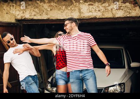 Eine Gruppe von zwei Jungen und ein Mädchen, lächeln und Spaß neben einem Parkplatz weißes Auto und runining Garage Stockfoto