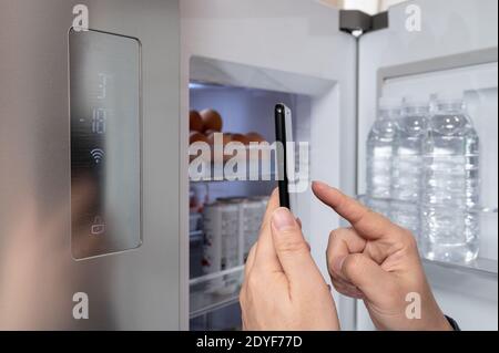 Die Hand des Mannes, der den Kühlschrank mit seinem Smartphone steuert. Internet der Dinge Konzept Stockfoto