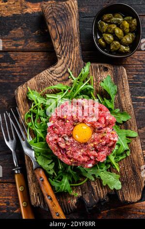 Auf einem Schneidebrett wird Tartar-Rind mit Wachtelei und Rucola serviert. Dunkler Holzhintergrund. Draufsicht Stockfoto