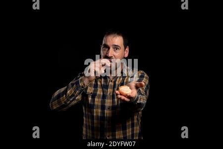 Bärtiger Mann in gelbem Karomuster, der mit einer Hand eine Orange anbietet und mit der anderen Hand auf schwarzem Studiohintergrund eine Scheibe isst. Gesund Stockfoto