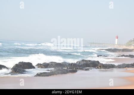Am frühen Morgen am Strand krachten Wellen gegen die Felsen mit Pier und Leuchtturm im Hintergrund Stockfoto