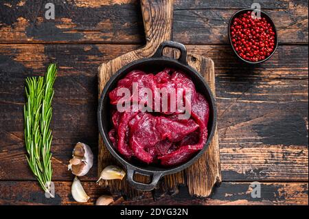 Frische rohe gewürfelte Scheiben von rotem Rinderfilet in gusseiserner Pfanne. Dunkler Holzhintergrund. Draufsicht Stockfoto