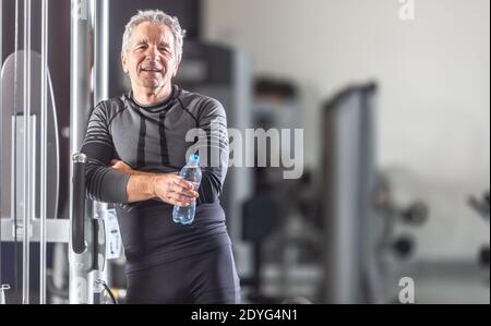 Ein alter Mann lehnt sich mit einer Flasche Wasser in den Händen an ein Trainingsgerät in einem Fitnessstudio. Stockfoto