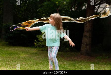Ein kleines Kind, kleines Mädchen, das riesige Seifenblasen im Garten macht. Aktives Kind im Hinterhof, grünen Park weht große gigante Blasen, Spaß haben Stockfoto