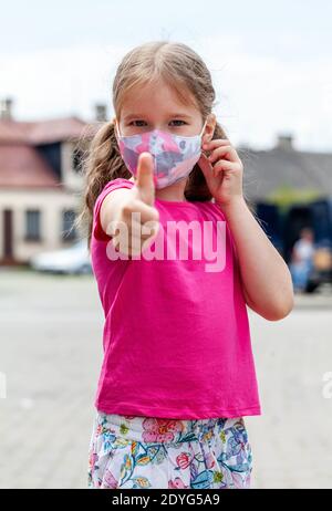 Kleines Mädchen, junges Schulkind trägt eine bunte Schutzmaske Gesicht zeigt Daumen nach oben Geste auf der Straße, Porträt, Nahaufnahme Stockfoto