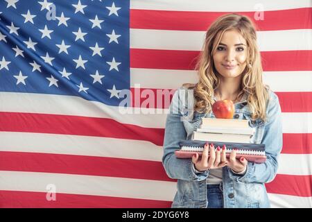 Junge Teenager-Studentin hält einen Stapel von Studienarbeiten und Apfel auf der Oberseite mit einer Flagge der Vereinigten Staaten hinter ihr. Stockfoto